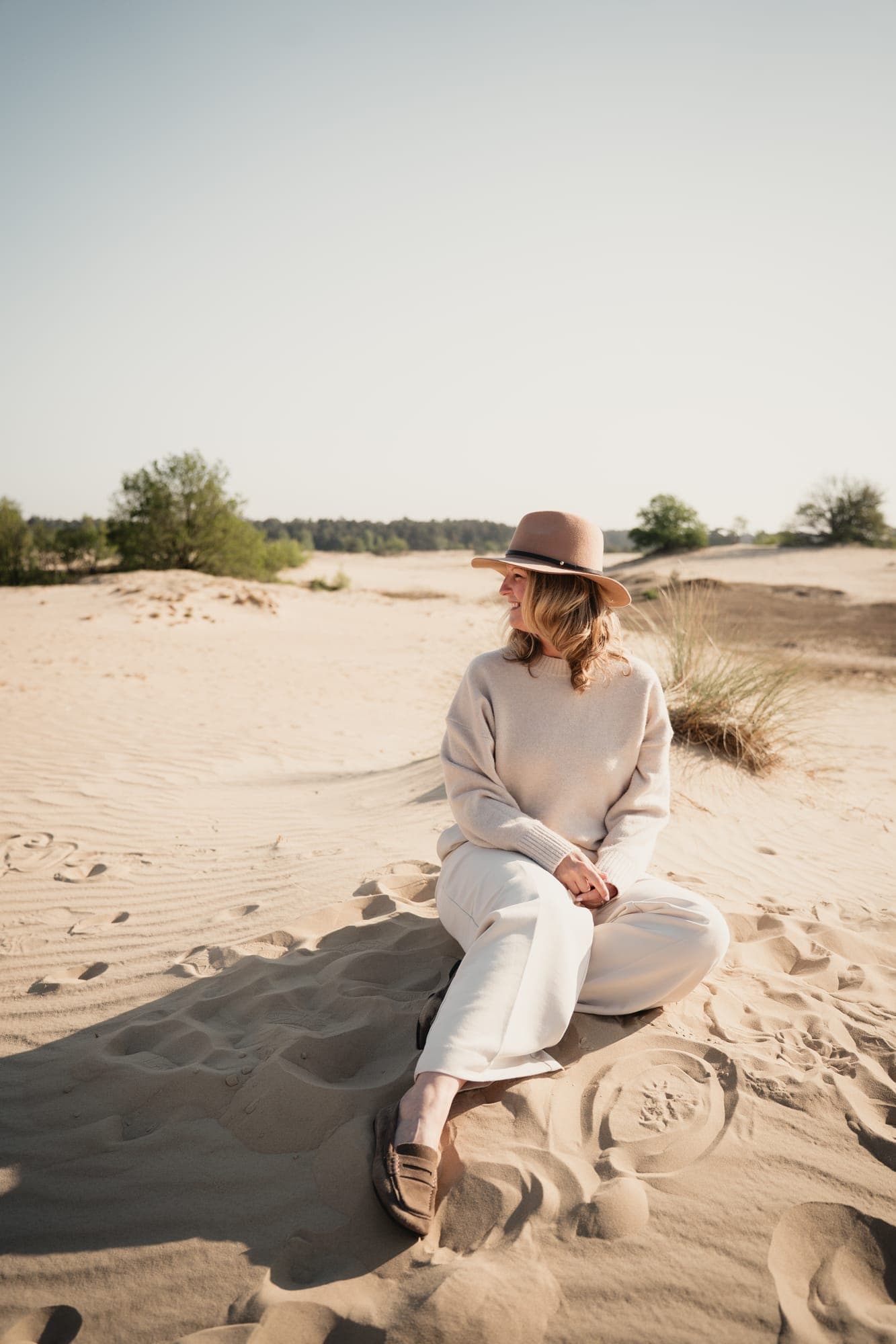 Een vrouw met hoed zit lachend in het zand van de duinen