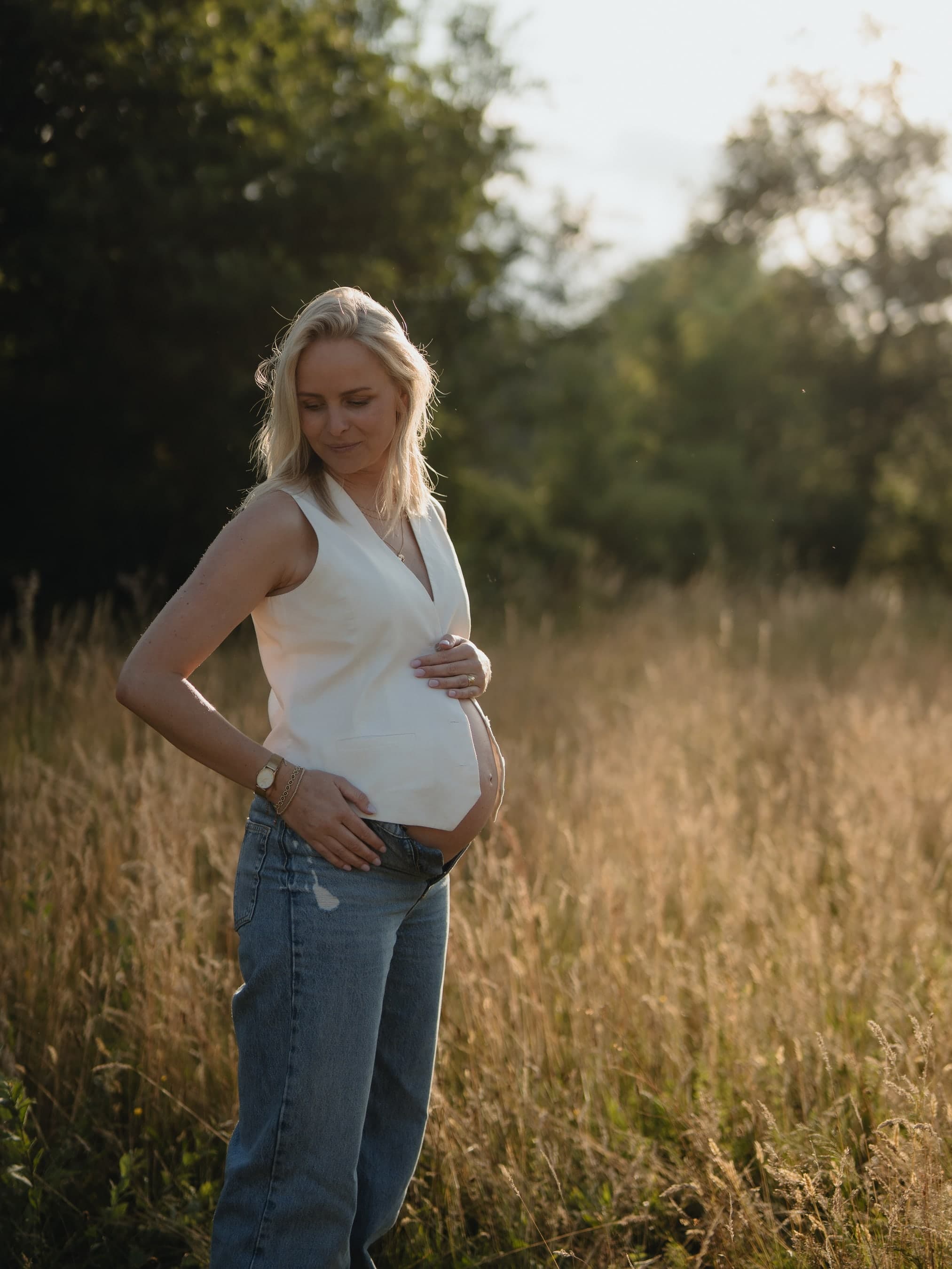 Een zwangere vrouw staat in hoog gras in warm avondlicht en kijkt glimlachend naar haar buik